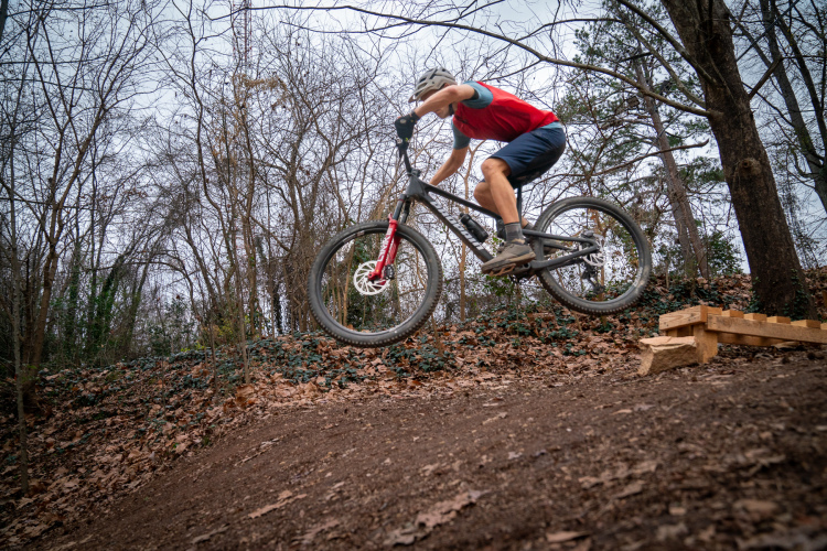 A cyclist performing a jump on a mountain bike over a wooden ramp in a wooded area with bare trees and fallen leaves on the ground.