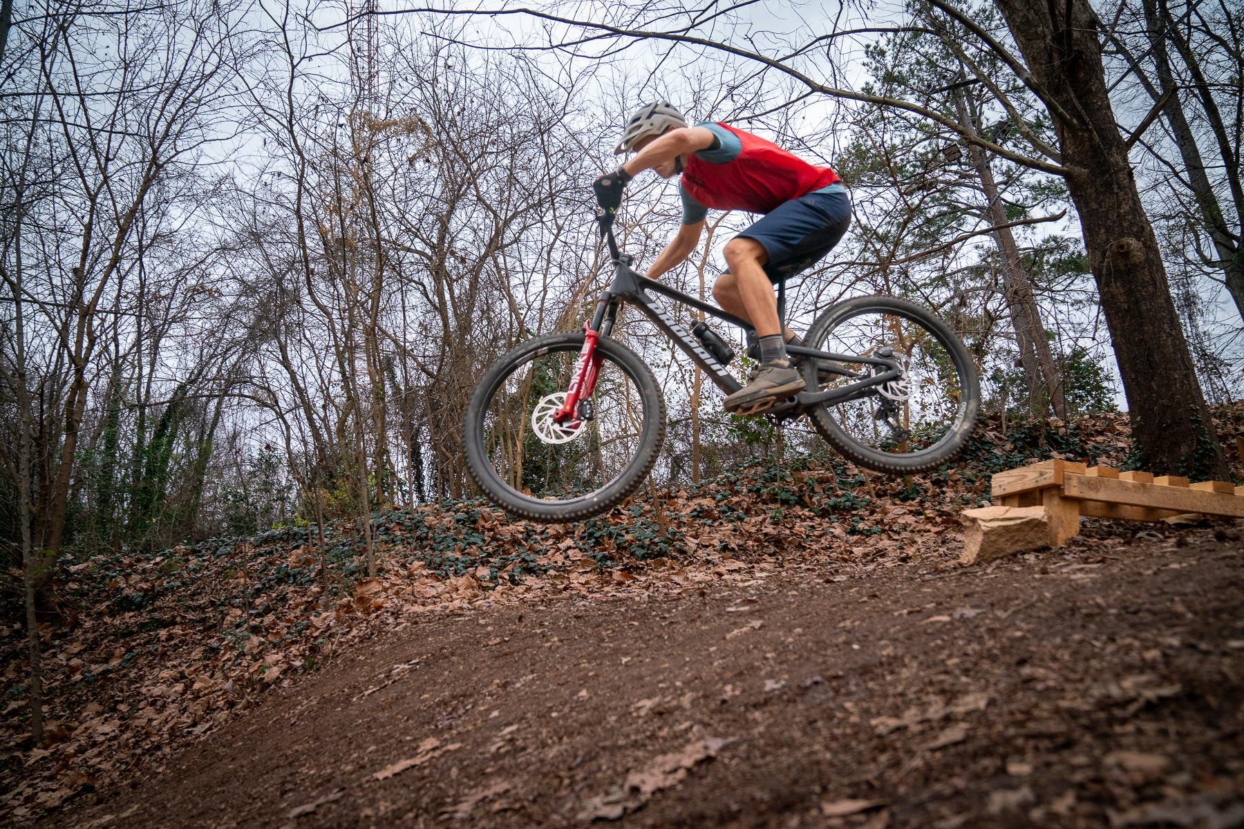 A cyclist performing a jump on a mountain bike over a wooden ramp in a wooded area with bare trees and fallen leaves on the ground.