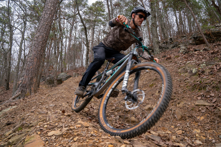 A mountain biker maneuvering down a rocky and wooded trail, leaning into the turn as they ride a black and teal bike with large tires. The scene features trees in the background and loose earth and leaves on the trail.