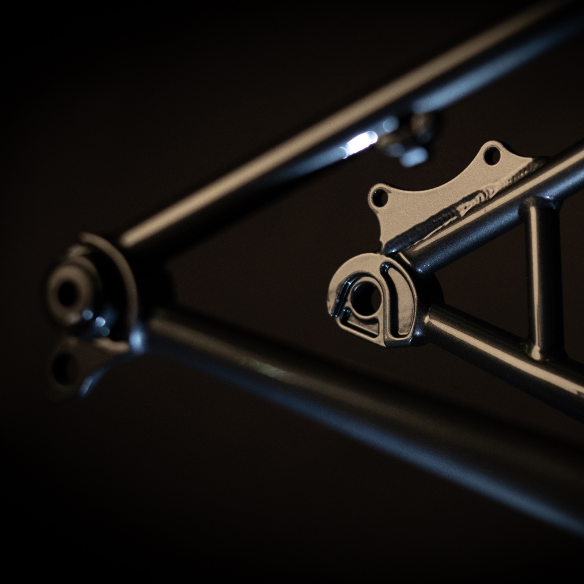 Close-up of a bike frame showing a detailed section with a metallic finish, highlighting the intricate design and structure of the tubing and junctions against a dark background.
