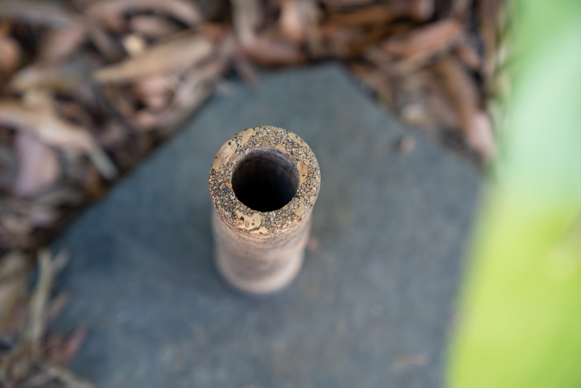 A close-up view of the top of a rustic, tapered pipe or tube, resting on a textured surface surrounded by dried leaves. The pipe has a slightly rough exterior with specks of dirt around its opening.