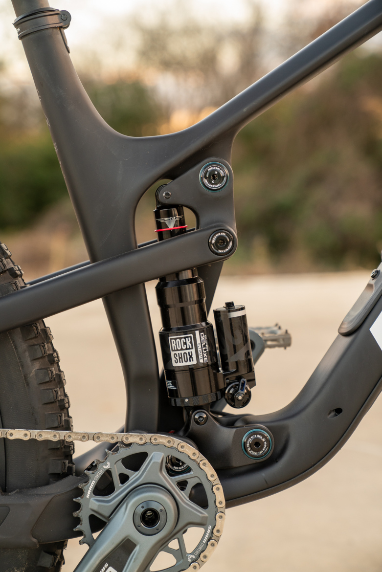 Close-up of a mountain bike's rear suspension system, showcasing a RockShox Deluxe shock and the bike's aluminum frame detailing. The image highlights the chainring, chain, and various components of the bike's drivetrain, with a blurred natural background.