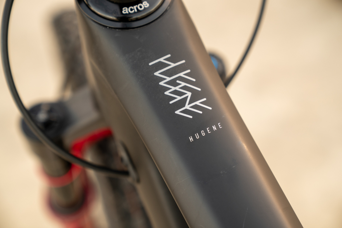 Close-up of a mountain bike frame featuring the logo "HUGENE" prominently displayed. The matte black finish of the frame contrasts with the white logo, while cable elements and other bike components are slightly blurred in the background.