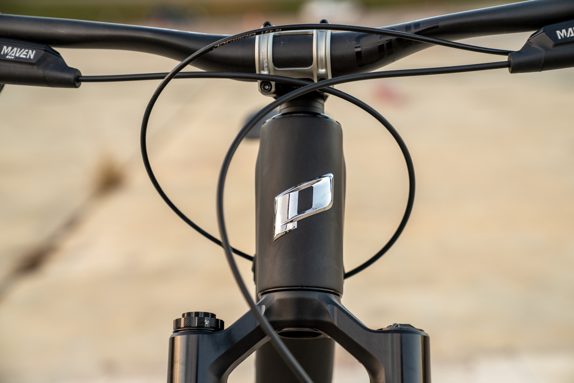 Close-up image of a bike's handlebar and stem, featuring a black frame and cables. The bike showcases a unique logo on the stem, with a background of a blurred outdoor setting.