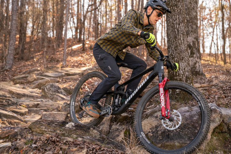 A mountain biker navigating rocky terrain in a forested area, wearing a helmet, sunglasses, and a plaid shirt, demonstrating skill and focus while riding a black mountain bike with red front suspension.
