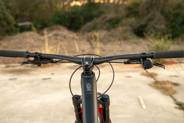 Close-up view of a mountain bike's handlebars, featuring grips, brake levers, and a gear shifter. The bike is positioned on a concrete surface with a natural background of dried grass and foliage.