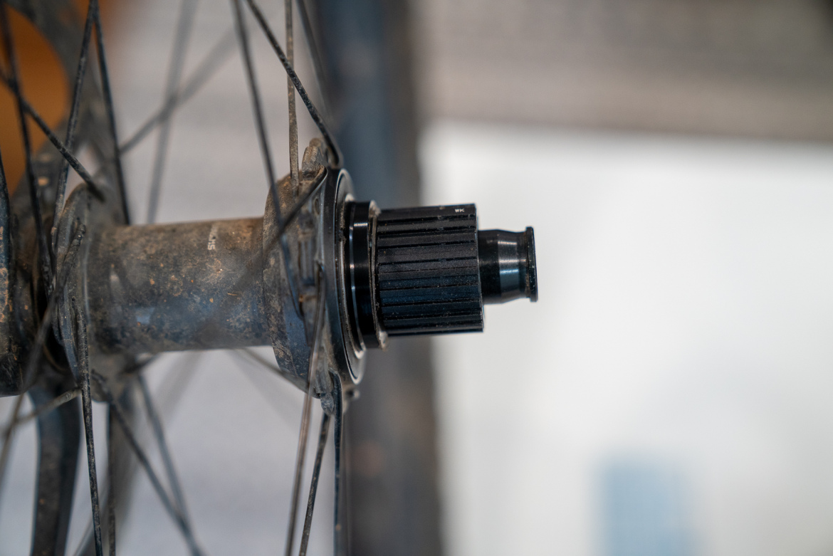 Close-up image of a bicycle wheel hub, showcasing the black freehub body attached to the hub shell. The hub shows signs of dirt, and the background features blurred elements that suggest a garage or workshop setting.