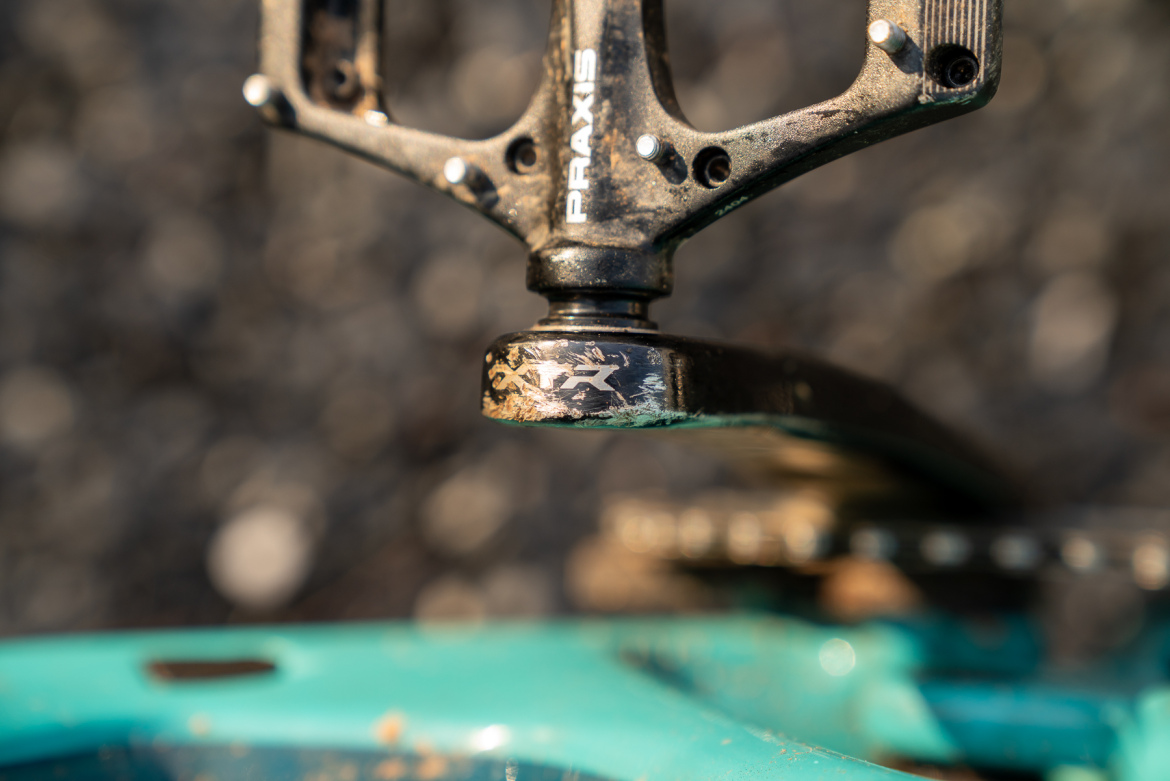 Close-up of a bicycle pedal with visible dirt and wear, attached to a crank arm. The bike frame is turquoise, and the background is a blurred mix of natural textures.
