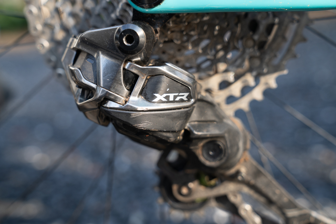 Close-up of a bicycle's rear derailleur, featuring a silver and black Shimano XTR component, with a background of a metallic bike cassette and gravel surface.