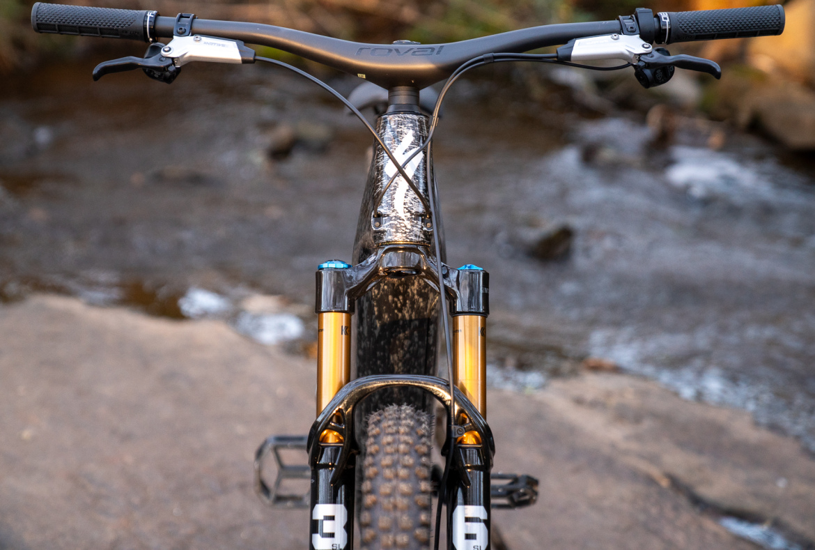 A close-up view of a mountain bike from the rear, showcasing the handlebars, front suspension forks, and part of the frame. The bike is positioned beside a flowing stream with natural rocky terrain in the background.