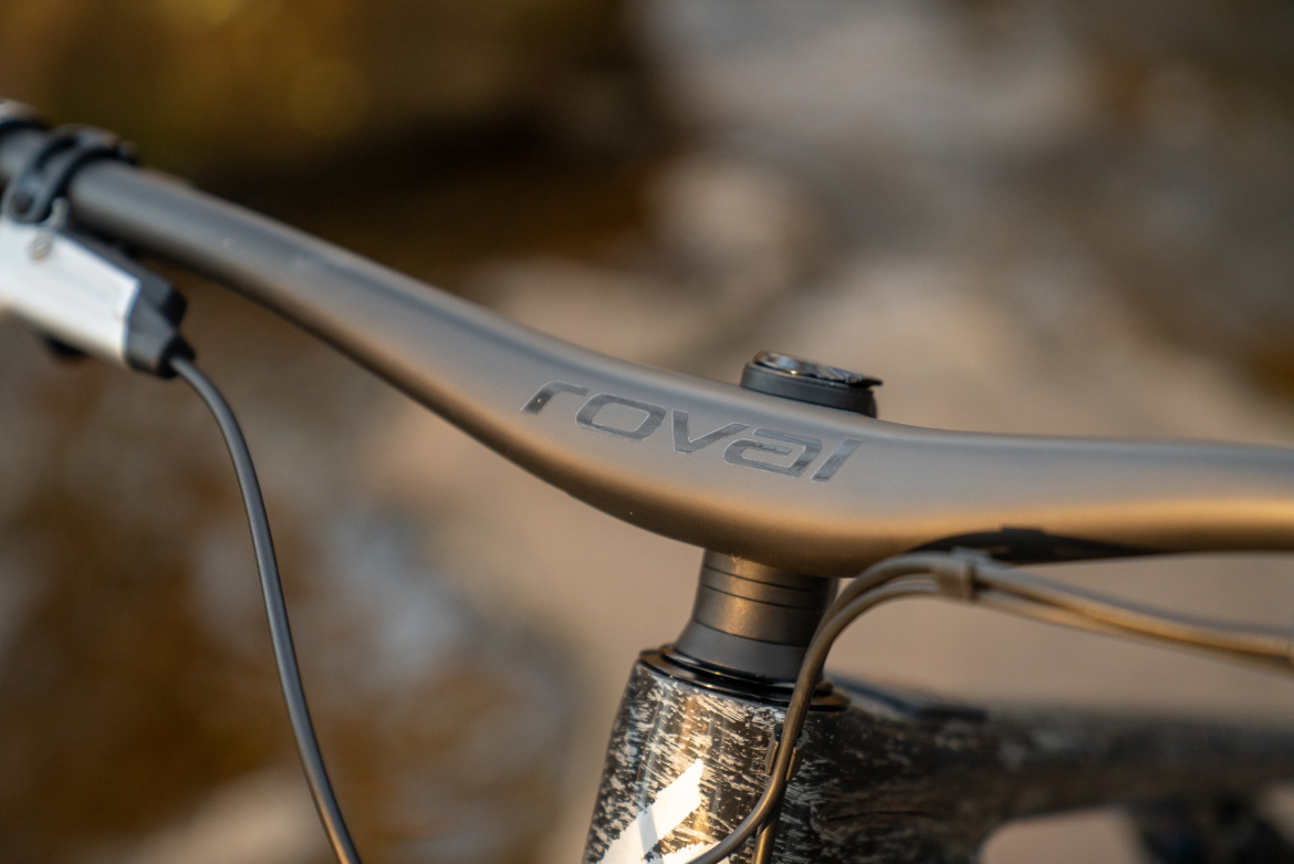 Close-up view of a bicycle handlebar featuring a sleek, black Roval brand design, showing the handlebar stem and cables. The background appears blurred, suggesting an outdoor setting.
