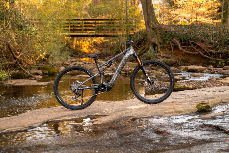A mountain bike resting on a rocky surface beside a calm stream, surrounded by trees with green foliage and autumn colors in the background. A wooden bridge can be seen in the distance, partially illuminated by warm sunlight.