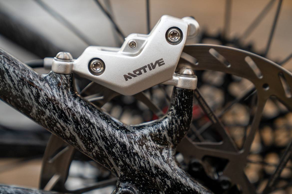 Close-up image of a silver bicycle brake caliper mounted on a black and white speckled bike frame, with a blurred background featuring a bicycle wheel and rotor.