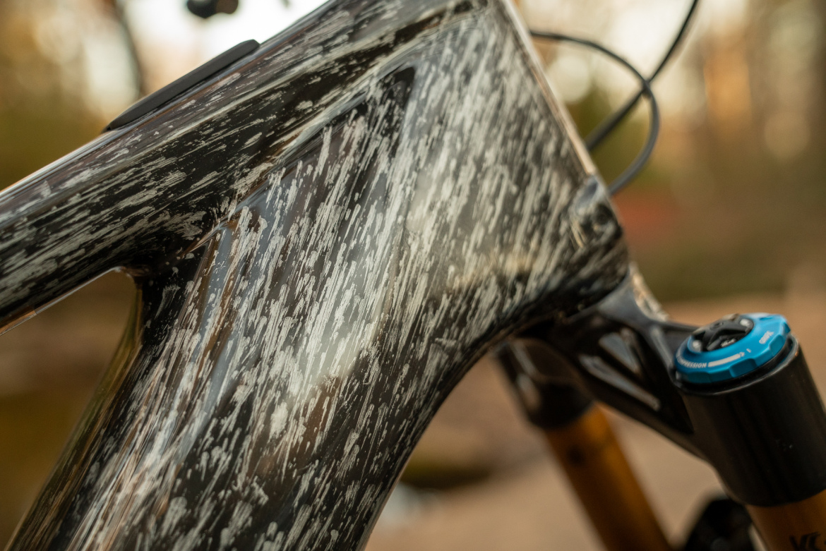 Close-up of a textured bicycle frame showcasing a black and white finish with a scratch-like pattern, highlighting its sleek design and construction details. The image also features a suspension adjustment dial in blue and gold components in the background, emphasizing the bike's high-performance features.