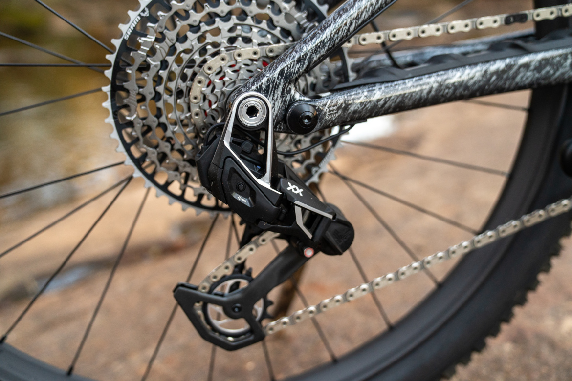 Close-up of a bicycle's rear derailleur, showcasing detailed components including the cassette, chain, and derailleur arm. The background features natural rock terrain and a blurred water element, emphasizing an outdoor cycling setting.