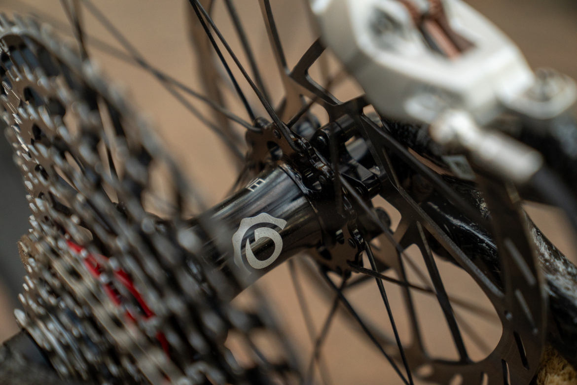 Close-up view of a bicycle's rear hub and cassette, featuring intricate metal components and a detailed logo on the hub. The background shows a blurred environment, emphasizing the bike's mechanics.