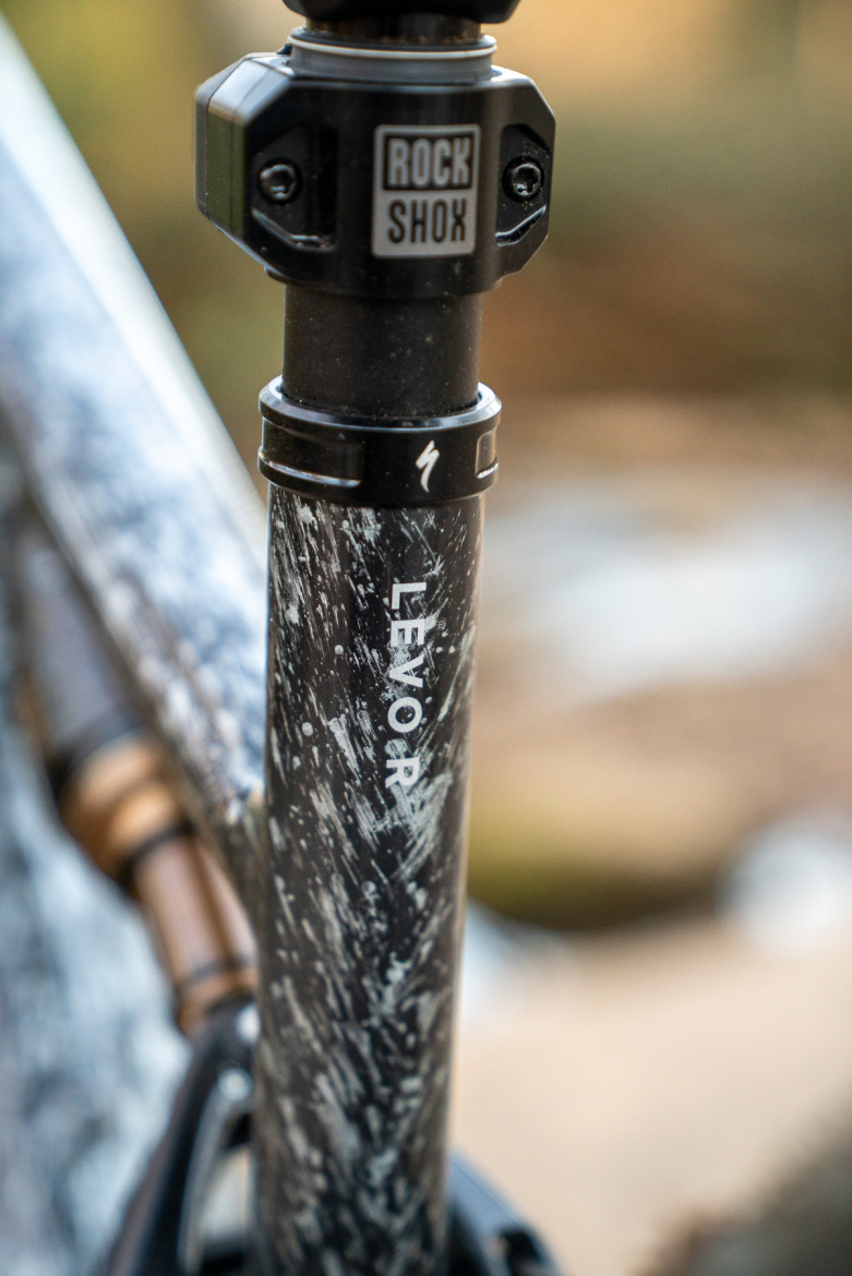 Close-up of a bicycle dropper post featuring the "LEVOR" label and a "Rock Shox" adjustable top assembly. The post has a textured black finish with some visible dirt splatter, indicating use in outdoor conditions.