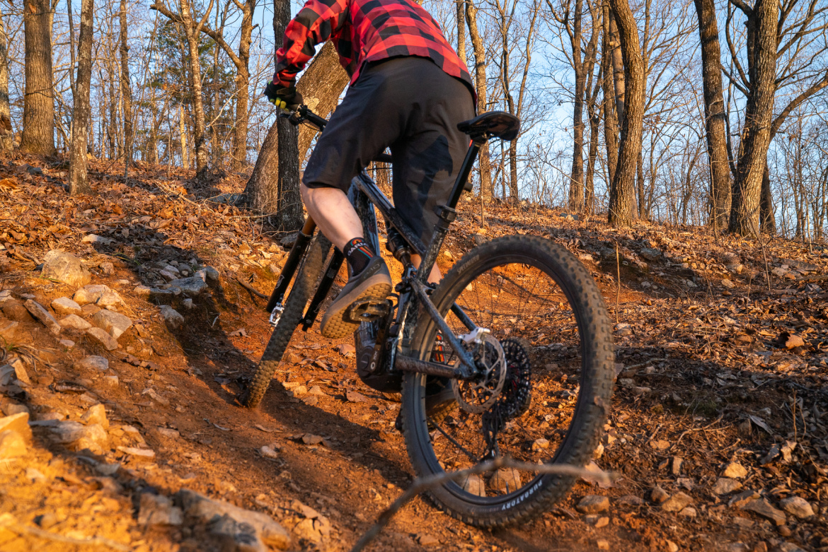 A cyclist wearing a red and black checkered shirt rides a mountain bike up a rocky, dirt trail surrounded by trees in a forest setting during sunset.