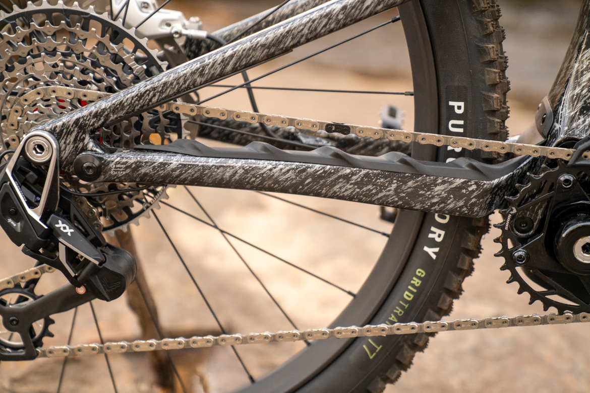 Close-up view of a modern mountain bike's drivetrain, featuring a carbon fiber chainstay, a visible gear cassette, and a derailleur, with a rugged tire in the background. The image highlights the intricate details of the bike’s components and chain.