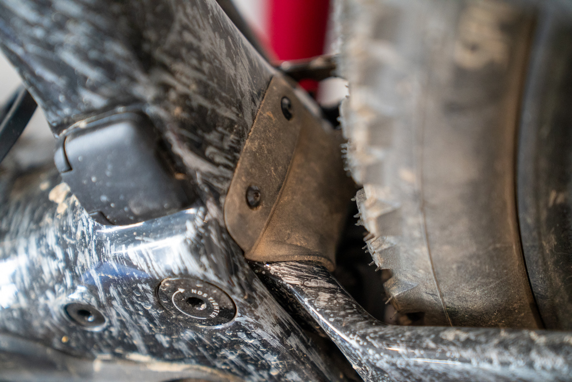 Close-up view of a mountain bike frame showing the lower section, with visible dirt and wear marks. The image highlights various components, including a protective rubber guard and part of the rear tire.