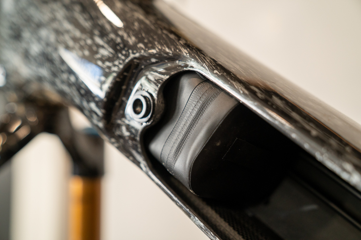 Close-up image of a bicycle frame showing a compartment with a black padded bag inside, featuring a textured surface and metallic accents. The frame has a glossy finish with a speckled pattern.