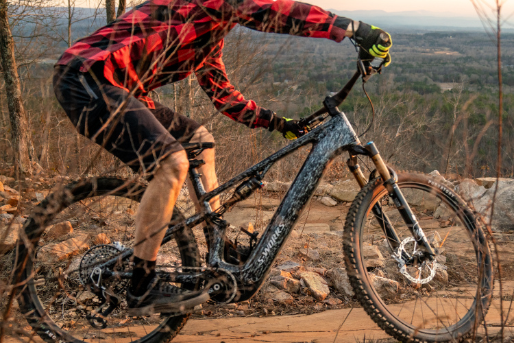 A cyclist in a red and black plaid shirt rides an electric mountain bike on a rocky trail, surrounded by sparse trees and hills in the background during sunset.