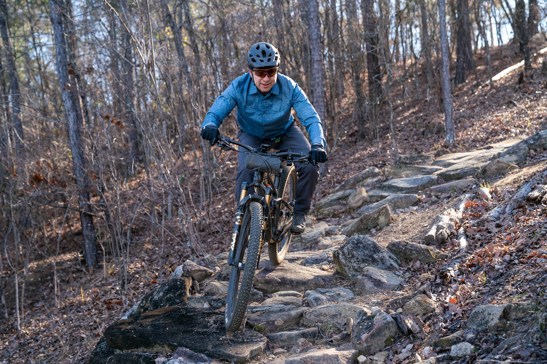 A mountain biker navigating through a rocky trail in a wooded area, wearing a helmet and sunglasses, with sunlight filtering through the trees. Standing Boy Trails mountain bike trail.