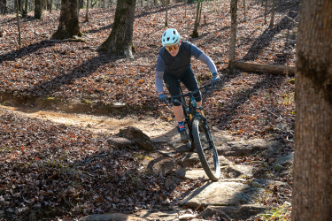 A person riding a mountain bike on a rocky trail in a forest, surrounded by trees and fallen leaves. The cyclist is wearing a helmet, sunglasses, and biking attire, focused on navigating the terrain. Sunlight filters through the trees, casting shadows on the ground. Standing Boy Trails mountain bike trail.