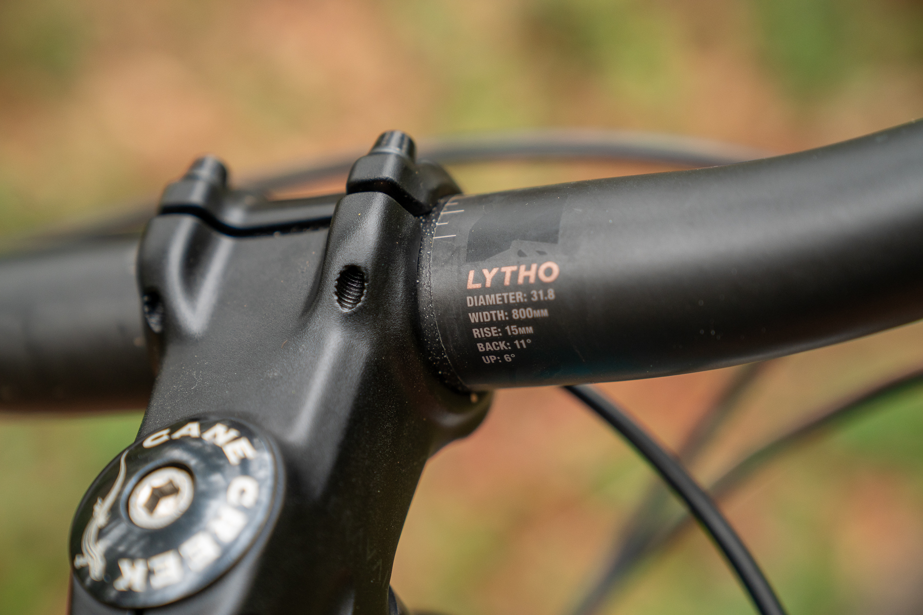 Close-up view of a bike handlebar stem displaying specifications, including brand name "LYTHO" and measurements for diameter, width, rise, back angle, and upward angle. The background features blurred natural elements, suggesting an outdoor setting.