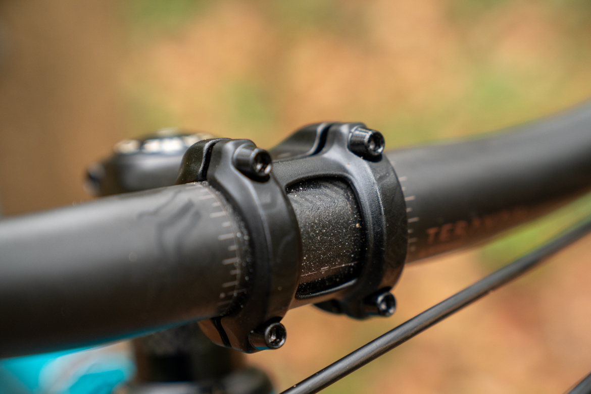 Close-up of a bicycle handlebar stem and grips, featuring black components with visible bolts and a slight layer of dust. The background is softly blurred, hinting at an outdoor setting with earthy colors.