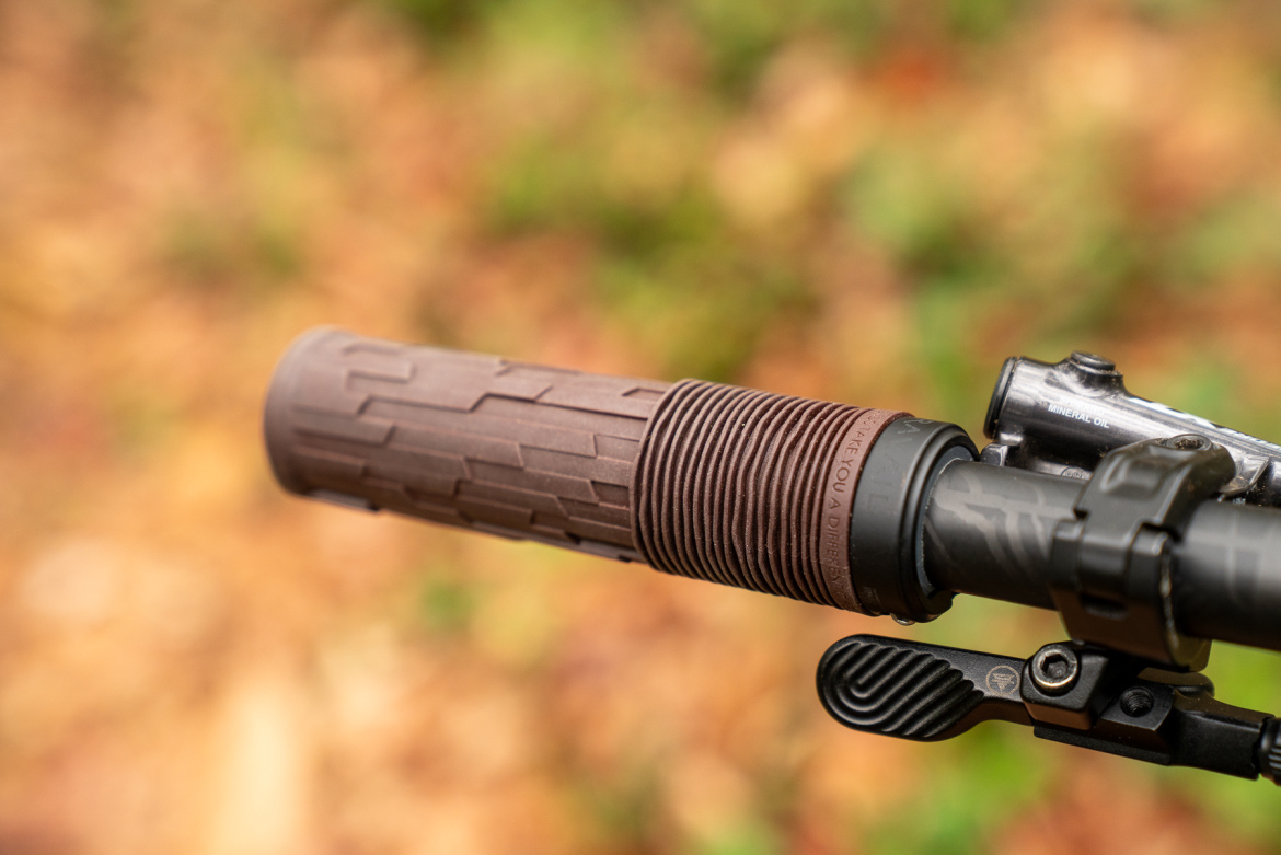 Close-up view of a textured bike grip on a handlebar, featuring a rubberized surface for better traction. The background shows a blurred natural setting with autumn foliage.