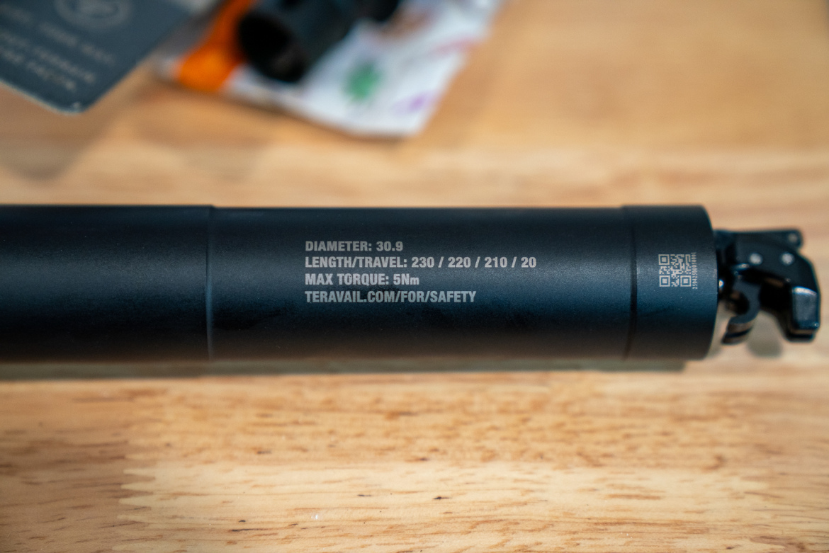 A close-up of a black bicycle seat post with labeled specifications, including diameter (30.9), length/travel measurements (230/220/210/20), maximum torque (5Nm), and a QR code. The post is resting on a wood surface, and background items are partially visible.