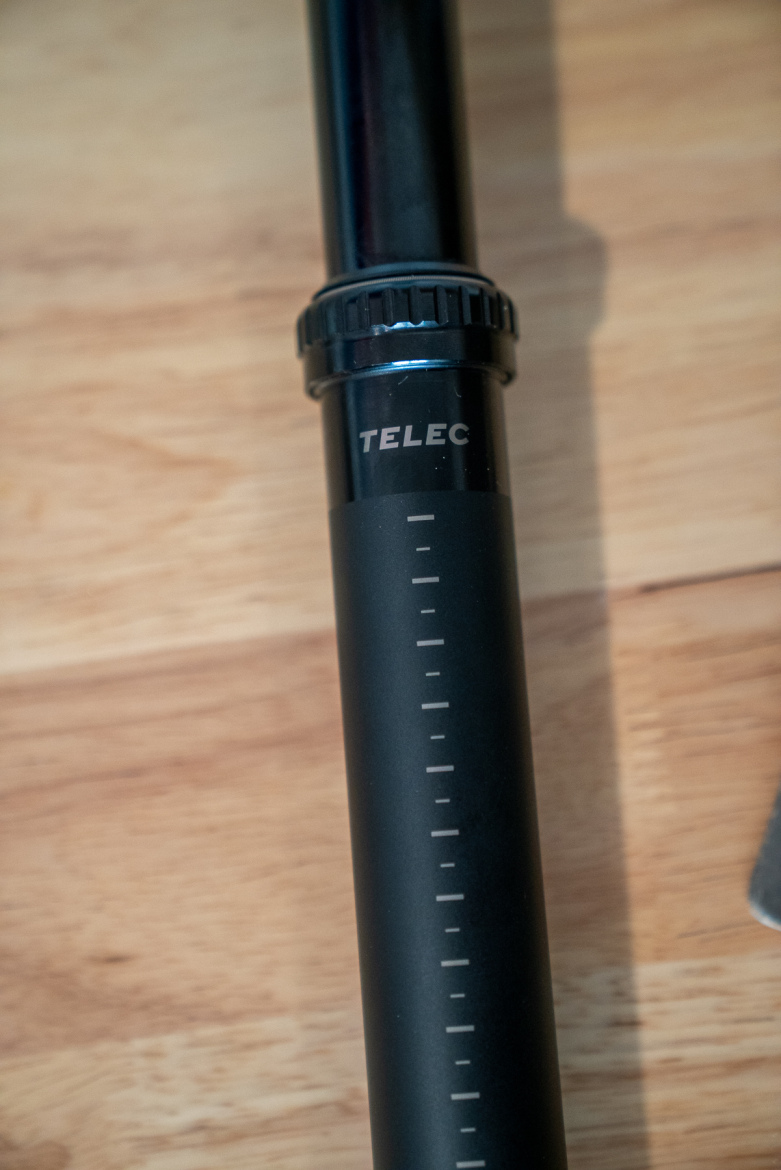 Close-up of a black telescopic pole with a measurement scale and the brand name "TELEC" visible. The pole is resting on a wooden surface.