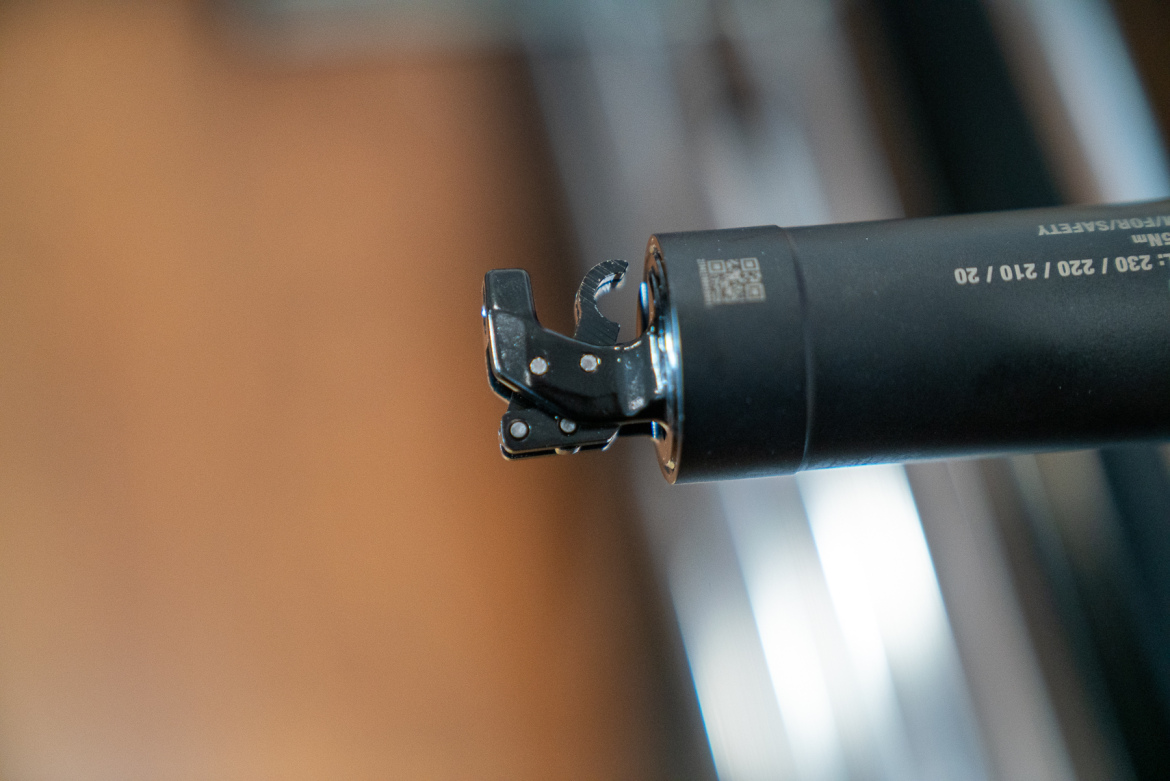 Close-up view of a mechanical component, featuring a black cylindrical body with a metallic lever and safety mechanism. The background is softly blurred, focusing attention on the part's details.