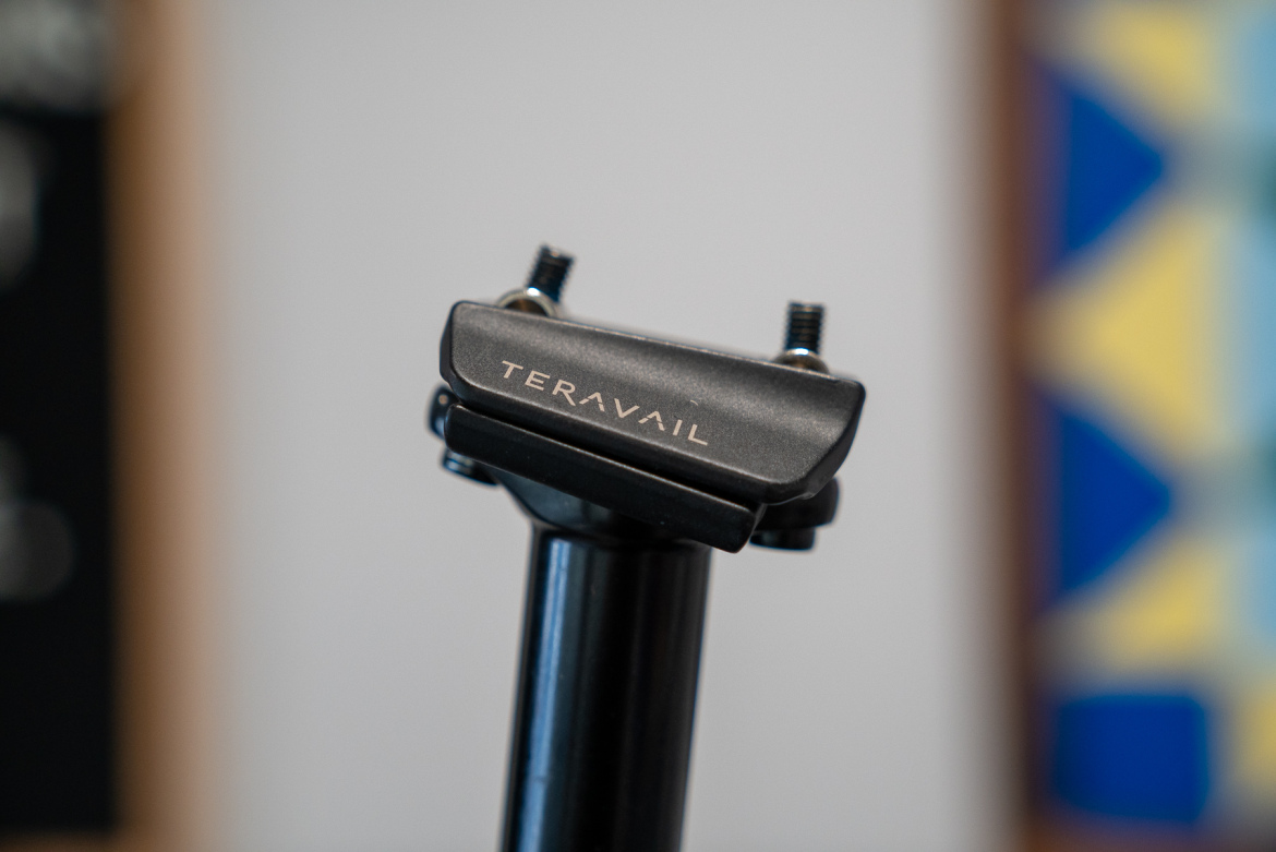 Close-up image of a black bike seat post with "TERAVAIL" engraved on the top portion. The post features two screws for adjustment and is set against a blurred background that includes geometric patterns.