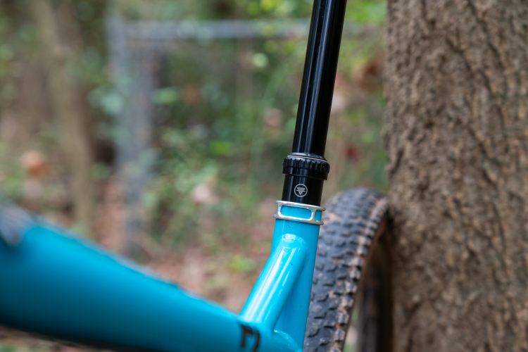 Close-up view of a mountain bike seat post, showcasing a turquoise frame and a black adjustable seat post clamp. The bike is positioned near a tree in a blurred outdoor setting, highlighting natural surroundings.