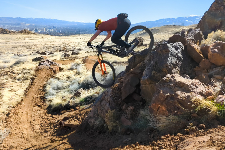 A mountain biker in a red shirt and yellow helmet is jumping off a rocky ledge on a dirt trail surrounded by dry grass and panoramic views of a valley and distant mountains.