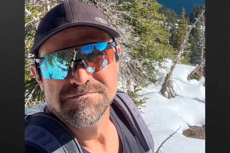 A person smiling in a winter landscape with snow-covered ground and evergreen trees in the background, wearing sunglasses and a backpack.
