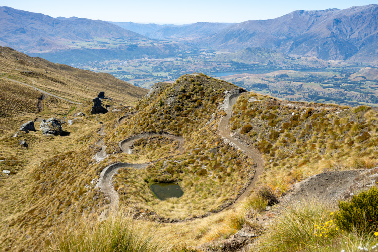 A panoramic view from a mountain ridge showcasing a winding dirt path that curves through grassy terrain, rocky outcrops, and a small pond, with rolling hills and valleys in the distance under a clear blue sky.