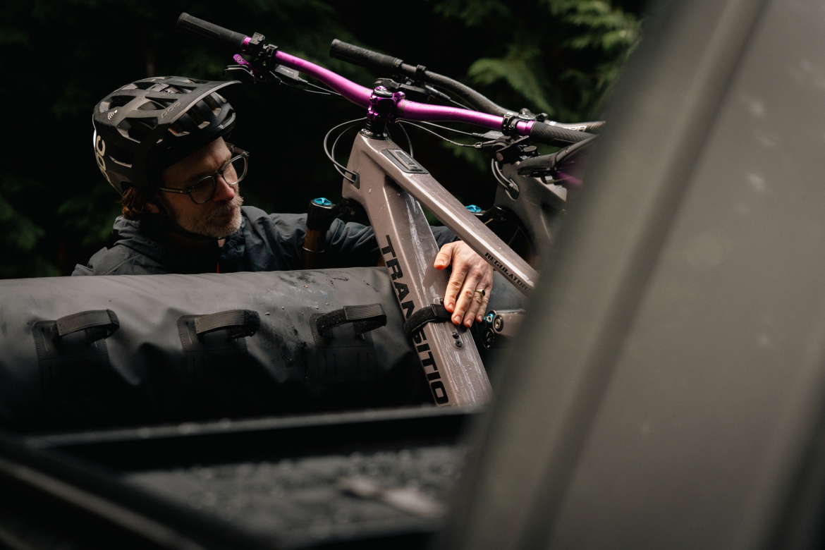 A person in a helmet and glasses is securing a mountain bike with a purple handlebar onto a vehicle. The background features dark green foliage, suggesting an outdoor setting, while the person is focused on adjusting the bike. A large black bag is also visible in the foreground.