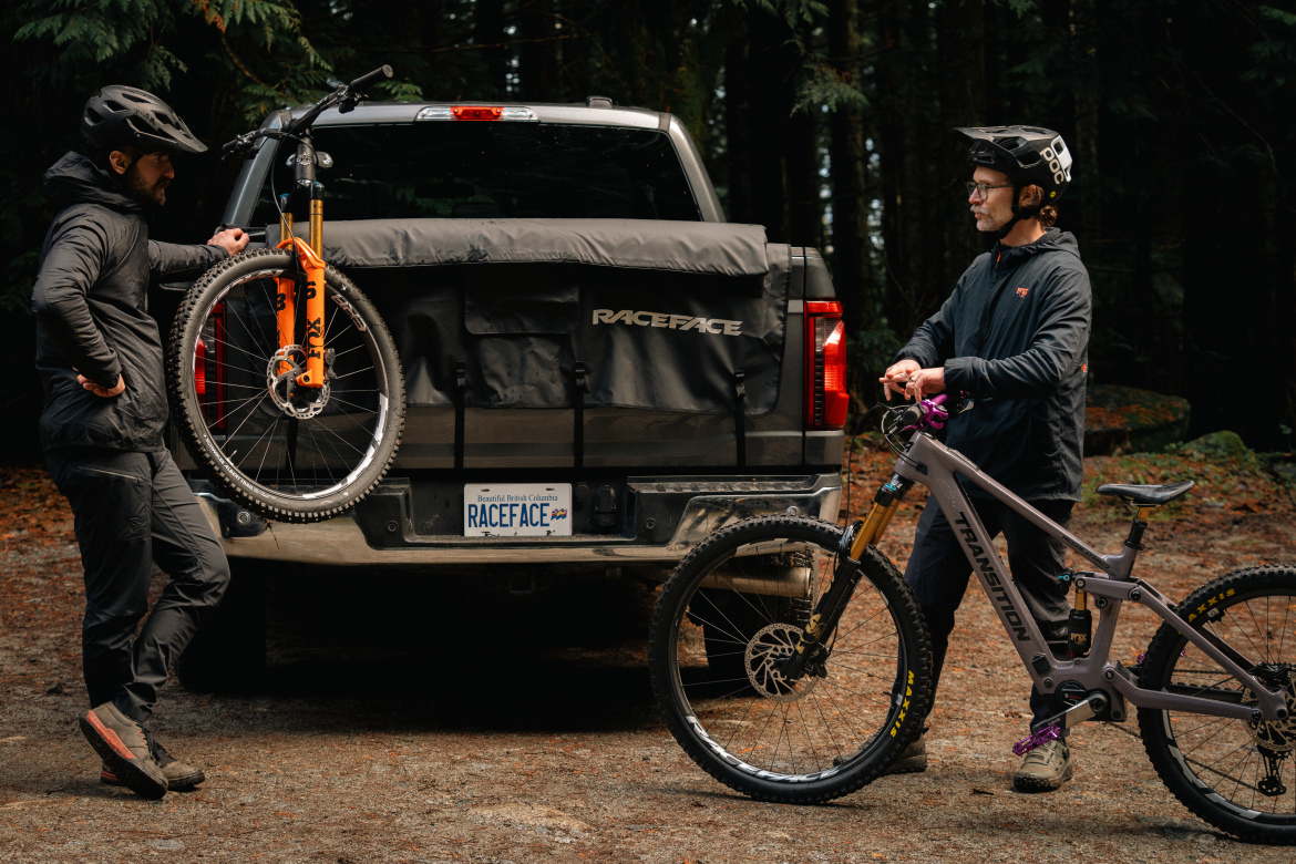 Two mountain bikers stand next to a pickup truck parked in a wooded area. One biker leans against the truck, which has a mountain bike with orange forks secured on its back. The other biker stands beside a gray mountain bike, holding it by the handlebars. Both wear helmets and casual outdoor clothing, and the surroundings are lush with trees and natural foliage.