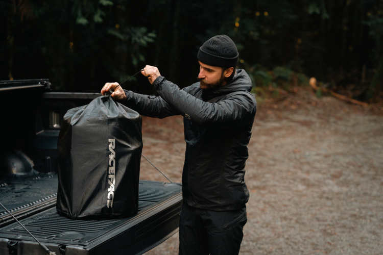 A person wearing a black jacket and beanie is standing near the open bed of a pickup truck, holding a black bag with the brand name "RACEFACE" visible. The background features a forested area with a gravel surface, suggesting an outdoor setting. The individual appears to be preparing or organizing their belongings.
