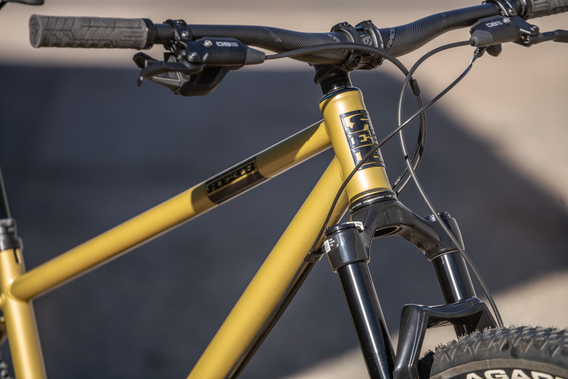 Close-up image of a mountain bike's handlebars and frame, featuring a matte yellow finish with black components. The handlebars are equipped with textured grips and gear shifters, while the frame prominently displays branding. The front fork shows a metallic finish, and the bike tire is partially visible in the bottom right corner.