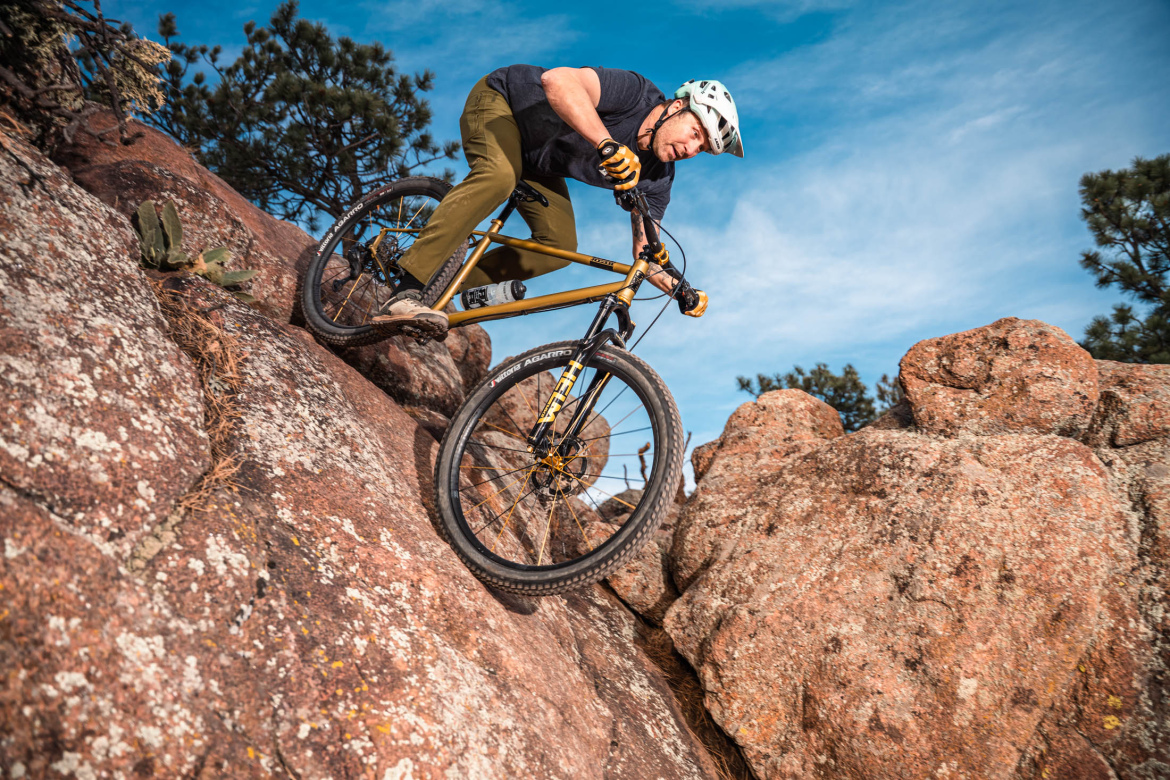 A mountain biker descending a rocky slope, wearing a helmet and riding a yellow mountain bike. The background features a blue sky with scattered clouds and pine trees among large boulders.