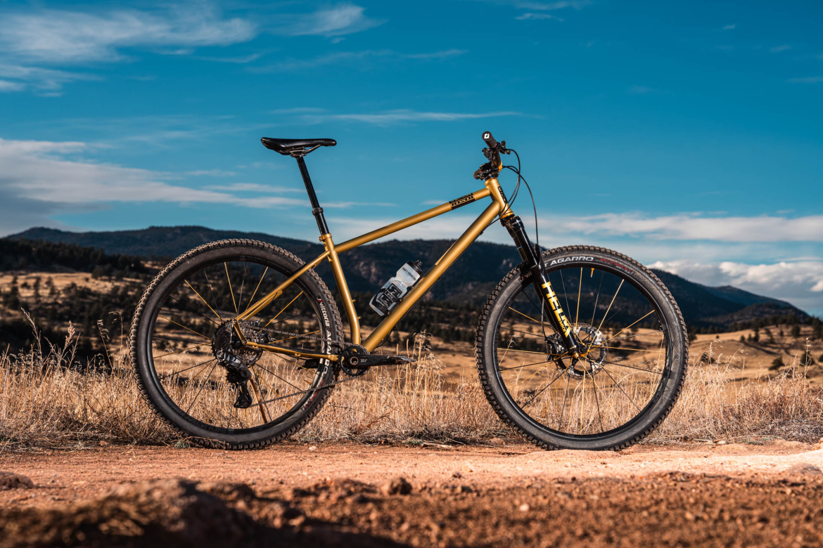 A gold mountain bike stands on a dirt trail surrounded by open fields and hills under a partly cloudy sky. The bike features thick tires and a water bottle mounted on the frame, highlighting its readiness for outdoor adventures.