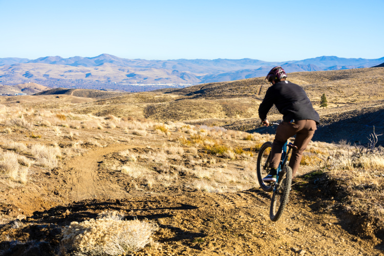 Mountain biker riding along a dirt trail in a hilly landscape, with dry grass and shrubs surrounding the path and distant mountains under a clear blue sky.