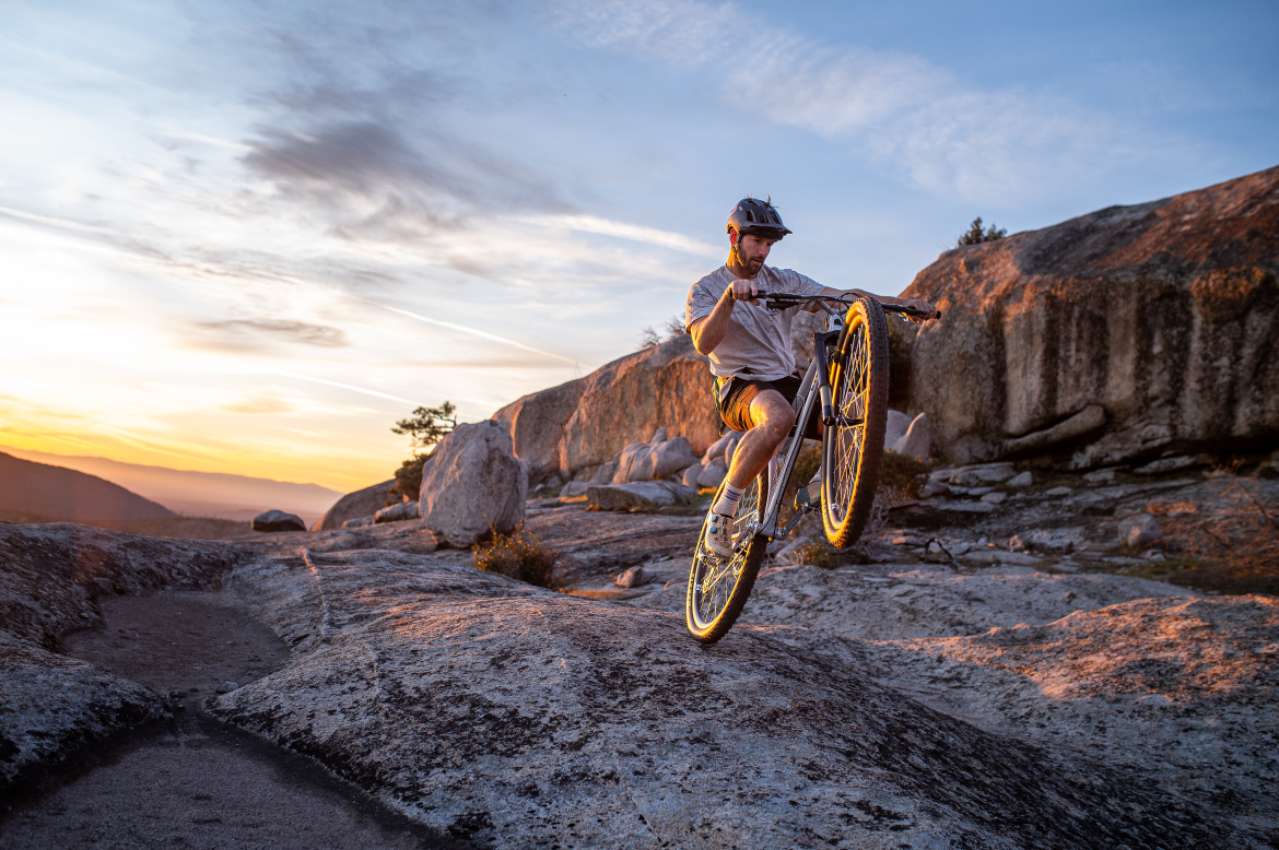 A mountain biker performing a wheelie on rocky terrain during sunset, with a scenic landscape in the background illuminated by warm light.