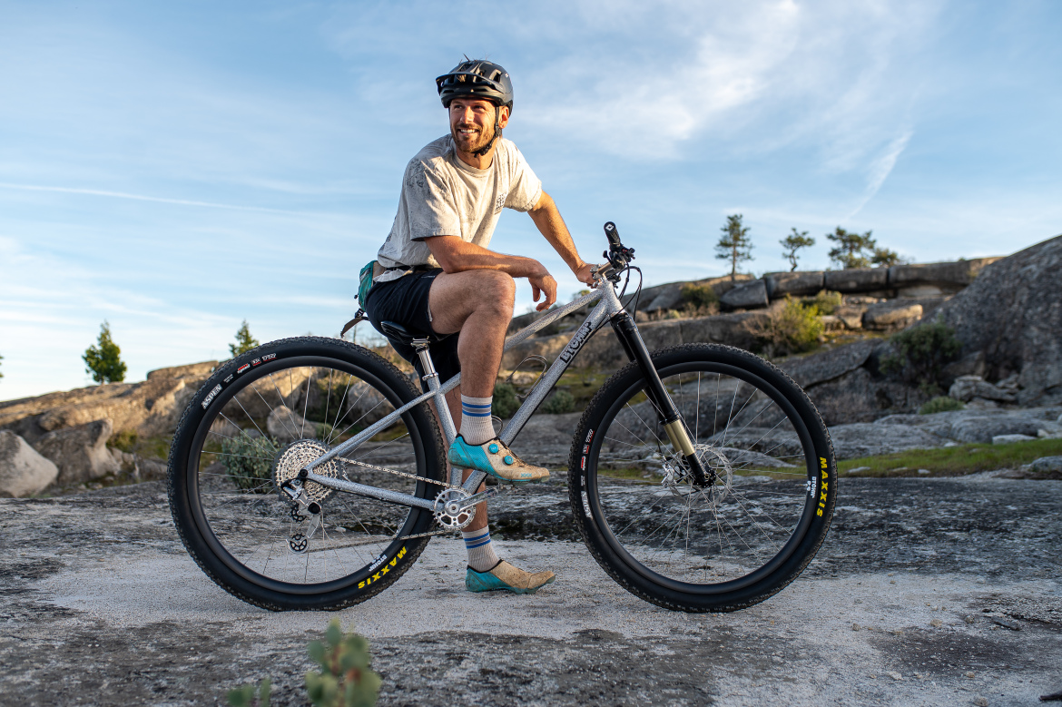 A smiling person sitting on a mountain bike, dressed in a gray t-shirt, shorts, and cycling shoes. The bike features large tires and is positioned on rocky terrain with a clear sky in the background. Trees are visible in the distance.