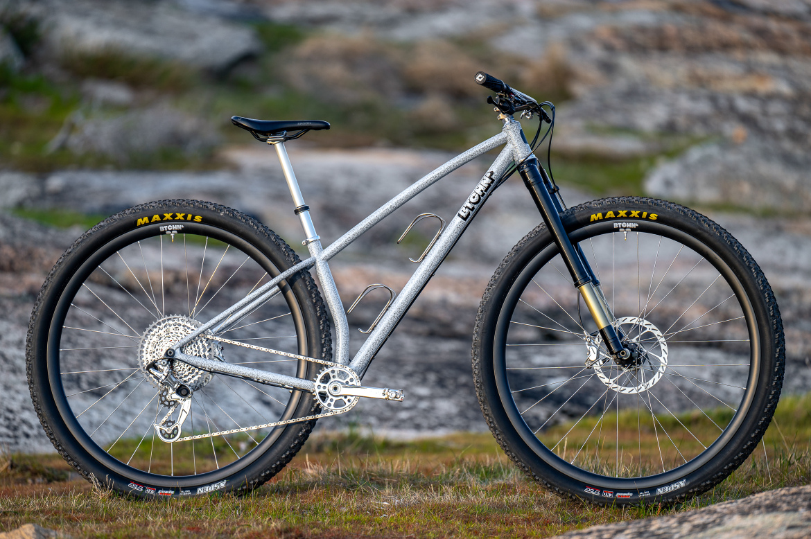 A silver mountain bike with a sleek frame, featuring black Maxxis tires and a disc brake system, is positioned against a rocky outdoor backdrop. The bike's design includes a prominent suspension fork and detailed gearing, highlighting its rugged appeal for off-road cycling. The ground around it is grassy with scattered rocks, suggesting a natural setting perfect for adventurous rides.