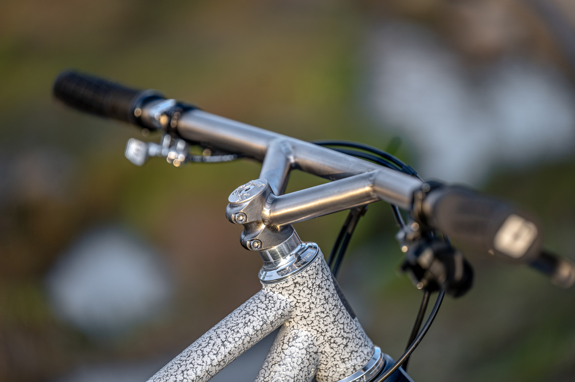 Close-up view of a bicycle handlebar and stem, featuring a textured gray finish on the frame. The handlebars are equipped with grips and brake levers, with cables neatly integrated. The background is softly blurred, highlighting the bike components.